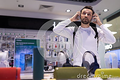 Photo of an adult handsome thinking brunet man with stubble in a white sweatshirt chooses headphones in an electronic Stock Photo