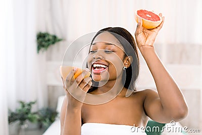 Photo of african american girl holding a half of grapefruit in front of her face and smiling on light background in spa Stock Photo