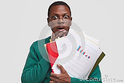 Photo of aghast stupefied dark skinned guy keeps hand on chest, analyzes information from paper documents, studies Stock Photo