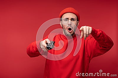 Photo of agitated man wih beard in red clothing pointing fingers at camera holds tv console with surprise and opened Stock Photo