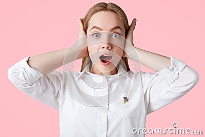 Photo of annoyed female plugs ears as hears loud music, has shocked expression, blue eyes and soft healthy skin, wears white blous Stock Photo