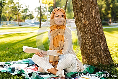 Photo of attractive islamic female student wearing headscarf sitting on blanket in green park Stock Photo