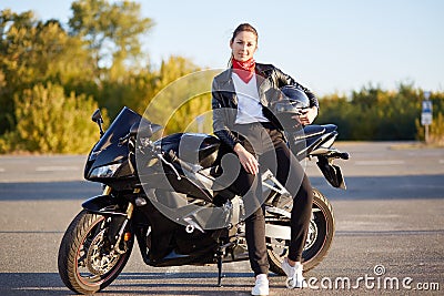 Photo of attractive slim female with ponitail, standing near black fast motobike with helmet in hand, wearing stylish outfit, has Stock Photo