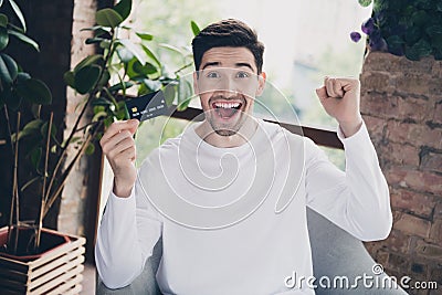 Photo of cheerful lucky young guy dressed white shirt rising fist showing bank card indoors house room Editorial Stock Photo