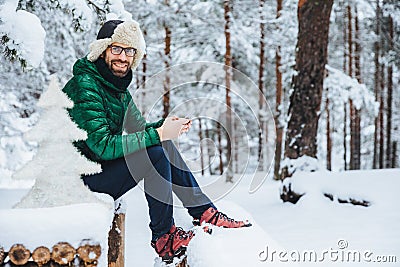 Photo of cheerful male types messages on smart phone looks directly into camera, sits in white winter forest, smiles happily, enjo Stock Photo