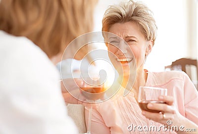 Cheerful mature friends laughing at old times while drinking red wine at home with yellow lens flare in background Stock Photo