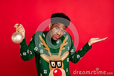 Photo of clueless unsure guy hold ball toy shrug shoulders wear green deer sweater isolated on red background Stock Photo