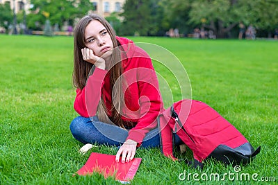 Photo of depressed passive sad teen girl sitting on green grass lawn is not willing to do home task looking away leaning on hand Stock Photo