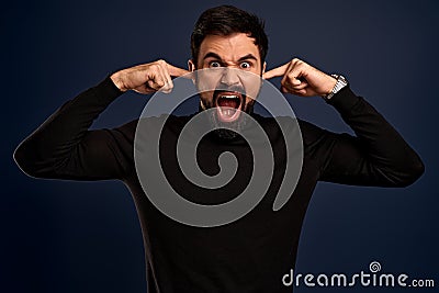 Photo of desperate annoyed bearded man keeps hands on head, shouts loudly, closes ears with his fingers, opens mouth, closes eyes Stock Photo