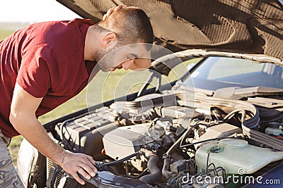 Photo of desperate man scratches head in bewilderment as stands in front of opened brocken car hood, can`t remove demage, looks fr Stock Photo