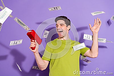 Photo of excited funky young man guy wear green t-shirt shooting money gun smiling isolated violet color background Stock Photo