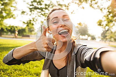 Photo of feminine fitness girl 20s in sportswear wearing bluetooth earpod taking selfie and showing thumb up, while walking in gr Stock Photo