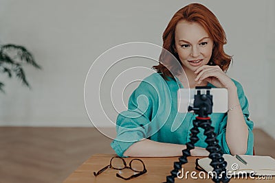 Photo of ginger female blogger shoots video of herself for personal blog, looks at camera of smartphone on tripod, chats with Stock Photo