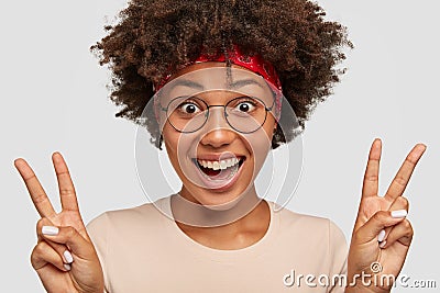 Photo of glad Afro American lady shows victory or peace sign, gestures with both hands, smiles broadly, wears round Stock Photo