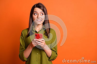 Photo of guilty young girl hold smartphone bite lip look side empty space wear green shirt isolated orange color Stock Photo