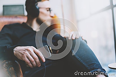 Photo handsome bearded man headphones listening to music modern loft studio.Man sitting in vintage chair looking window,holding sm Stock Photo