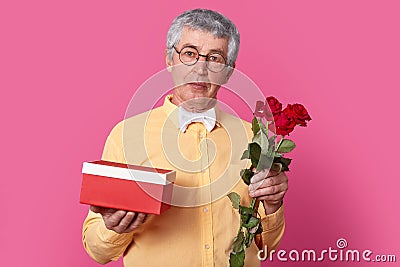 Photo of handsome of mature man in spectacles, dressed in yellow shirt with bowtie, carries red box with present and roses, wants Stock Photo