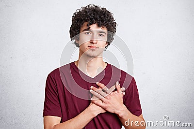 Photo of handsome serious male keeps hands on chest, wears casual t shirt, expresses his friendly feeling to people who surround h Stock Photo