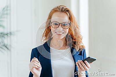 Photo of happy female leader with ginger hair, wears spectacles, updates app on modern cell phone, wears elegant clothes, poses in Stock Photo