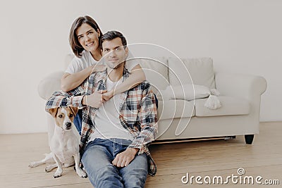 Photo of happy woman embraces her husband, poses on sofa, little dog sits on floor near owner, look all in camera with happy Stock Photo