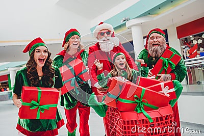 Photo of impressed amazed santa claus assistants wear costumes smiling riding cart buying many gift packages indoors Stock Photo