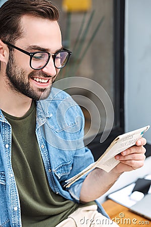 Photo of intelligent young man smiling and reading newspaper while sitting in city cafe outdoors Stock Photo