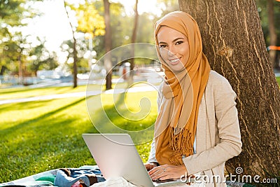 Photo of islamic female student 20s wearing headscarf sitting on blanket in green park Stock Photo