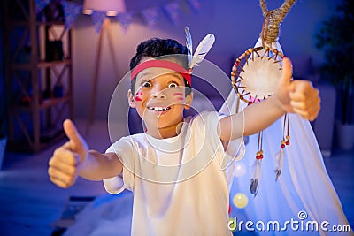 Photo of joyful surprised boy in native american costume have theme party birthday thumb up symbol in evening playroom Stock Photo