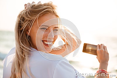 Photo of joyous blonde woman 20s smiling, and taking photo of sunrise above sea, while walking on summer beach Stock Photo