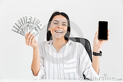 Photo of lucky businesswoman 30s holding smartphone and fan with dollar money banknotes while sitting in armchair in office Stock Photo