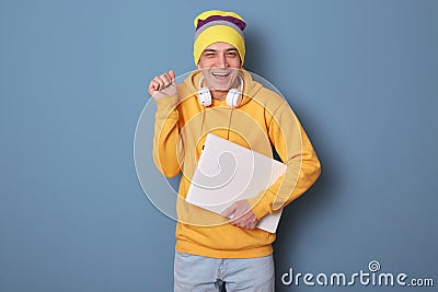 Photo of overjoyed smiling cheerful man wearing beanie hat and casual yellow hoodie, holding closed laptop, clenched fist, Stock Photo