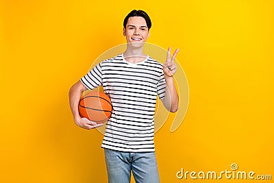 Photo of pleasant student dressed striped t-shirt hold bascketball ball showing v-sign symbol isolated on vivid yellow Stock Photo