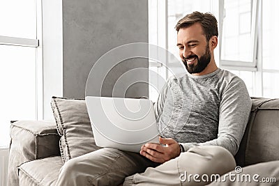 Photo of pleased adult man 30s in casual clothing typing on laptop, while working in cozy home Stock Photo