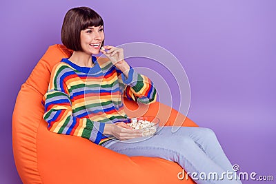 Photo of positive lady sit beanbag eat popcorn enjoy cinema wear striped pullover isolated violet color background Stock Photo