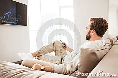 Photo of relaxed adult man 30s in casual clothing sitting on sofa in living room, and watching TV Stock Photo