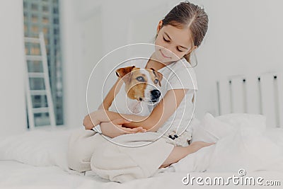 Photo of relaxed beautiful small girl plays with pedigree dog, embraces favourite animal, dressed in casual pyjamas, sits on bed, Stock Photo