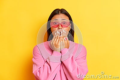 Photo of scared Asian woman dressed in pink hoodie and glasses grabs face and looks with worried expression she afraid Stock Photo