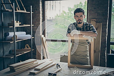 Photo of serious confident joiner in gloves uniform leaning on handmade frame looking at you pensively with planks and Stock Photo