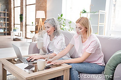 Photo of serious family grey haired mom and blonde daughter sit sofa write note use laptop indoors inside house home Stock Photo