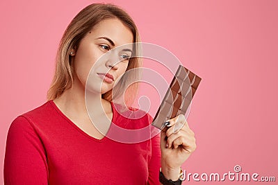 Photo of serious woman holds bar of cocoa chocolate, feels temptation, keeps to diet, dreessed in red outfit, isolated over pink Stock Photo