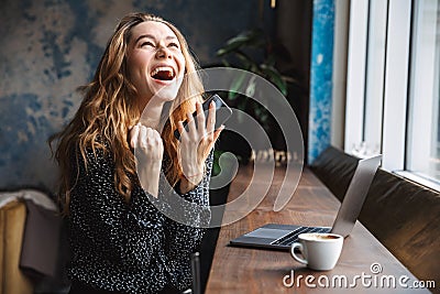 Excited beautiful young pretty woman sitting in cafe indoors using laptop computer and mobile phone Stock Photo