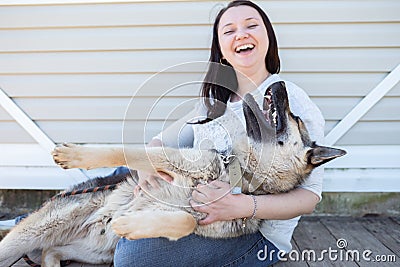 Photo of smiling woman in jeans and white jacket sitting next to happy shepherd puppy against white wooden building Stock Photo