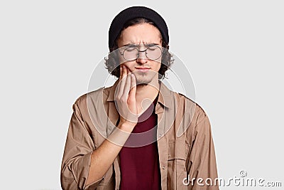 Photo of stressful youngster with small beard keeps hand on cheek, suffers from toothache, keeps eyes shut, dressed in stylish clo Stock Photo