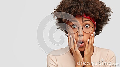 Photo of stupefied emotional black woman with bugged eyes, keeps hands on cheeks, stares through spectacles, models Stock Photo
