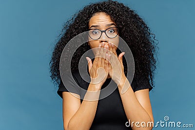 Photo of stupefied young female covers mouth, has bated breath, bugged eyes, Afro hair, cannot believe in shocking news, wears Stock Photo