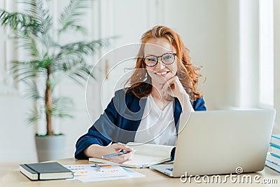 Photo of successful ginger female freelancer has remote work, watches webinar online on laptop computer, writes down information Stock Photo