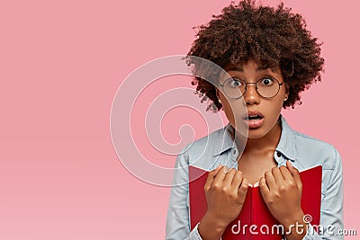 Photo of surprised scared dark skinned woman carries red textbook, reads something astonishing, gasps from fear Stock Photo