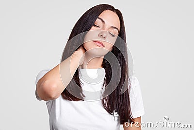 Photo of tired brunette female feels pain in neck as has sedentary lifestyle, needs physical activity, closes eyes, wants sleep, h Stock Photo