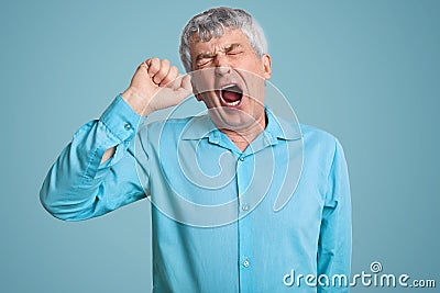 Photo of tired grey haired retired man feels sleepy, yawns as feels tired, opens mouth widely, dressed in elegant shirt, poses Stock Photo