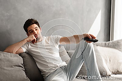 Photo of young bored man sitting on couch at home, and changing Stock Photo